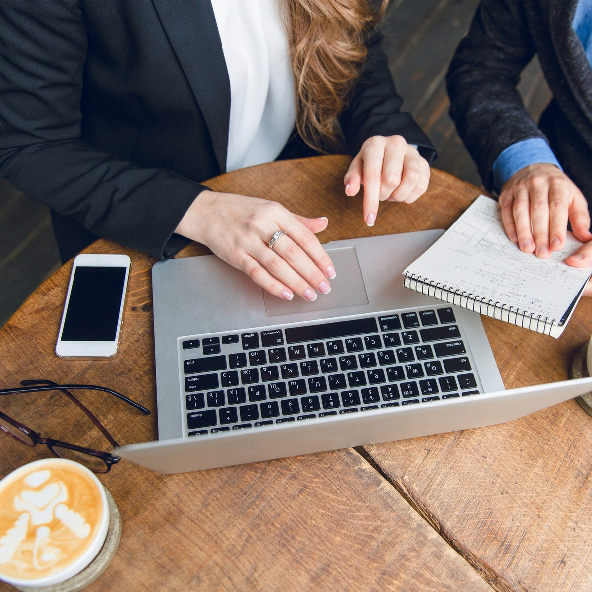 Dos personas trabajan en una mesa con laptop, libreta y café